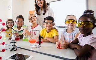 Students wearing safety goggles smiling at the table holding their beakers with multi colored liquid inside them.
