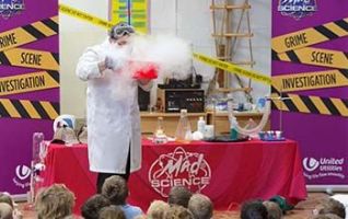 Mad scientist blowing into a red bowl with smoke coming out of it while kids watch